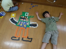 ドミノで作った作品の横に寝転ぶ児童の写真